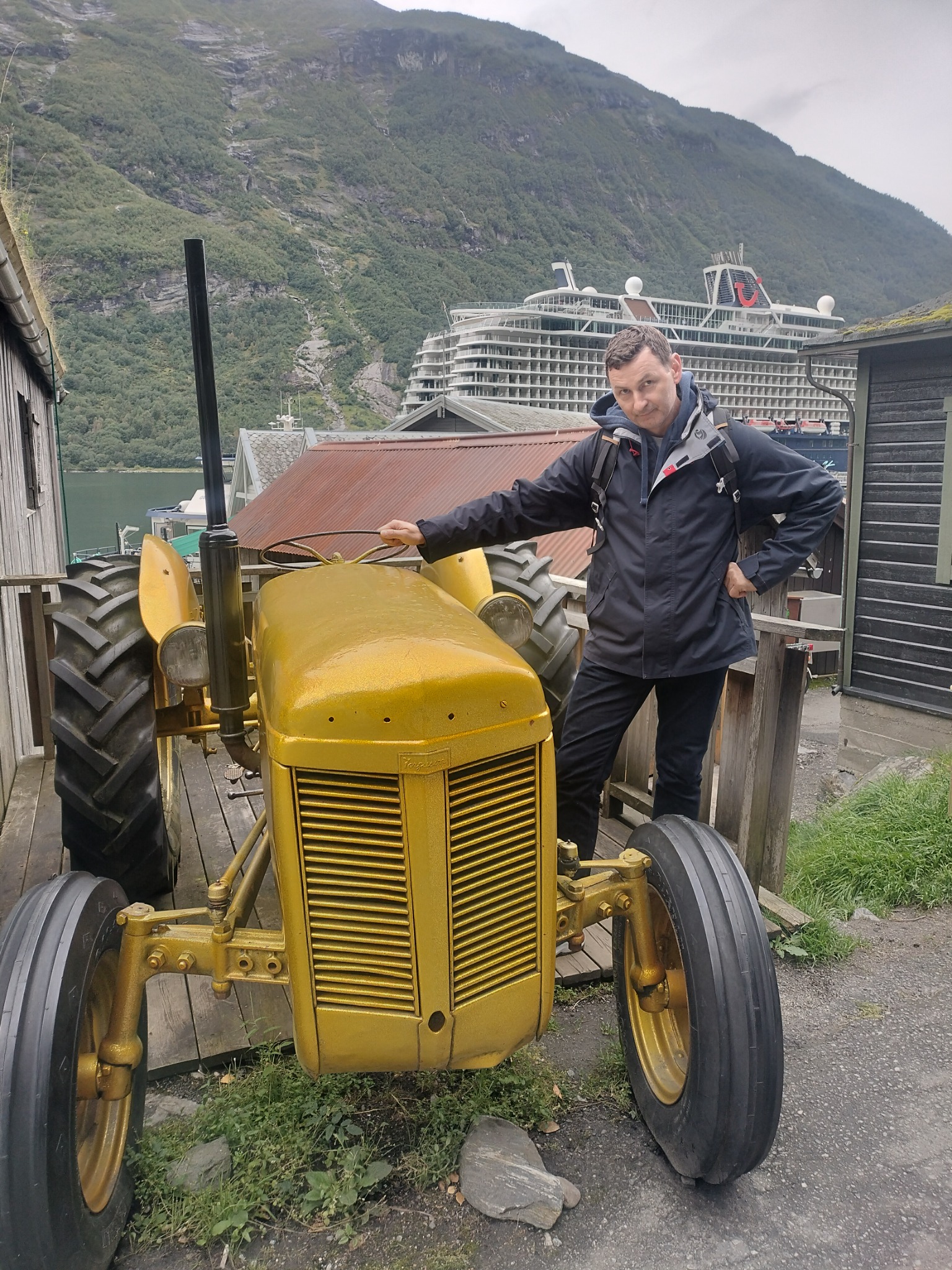 Mężczyzna pozuje obok odrestaurowanego, złotego traktora w scenerii norweskiego fiordu z dużym statkiem wycieczkowym w tle.