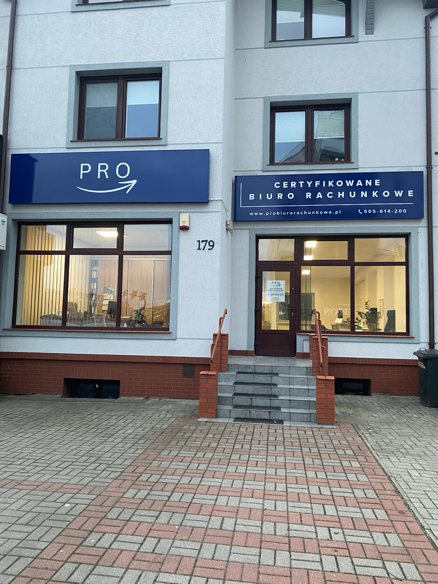 Fasada budynku biura rachunkowego z dwoma szyldami: 'PRO' z symbolem strzałki oraz 'CERTYFIKOWANE BIURO RACHUNKOWE' z adresem strony internetowej i numerem telefonu, widok z zewnątrz na wejście...