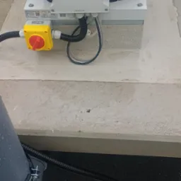 Widok z góry na zainstalowany wentylator dachowy Brookvent CSR-B-EC-W z żółtym wyłącznikiem, połączony przewodami w peszlach, na betonowej podstawie z widocznymi kroplami wody.