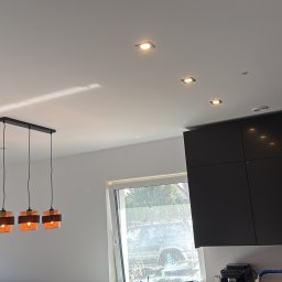 Zamontowane oświetlenie sufitowe: trzy lampy wiszące z pomarańczowymi kloszami i rząd punktowych halogenów w nowoczesnej kuchni z czarnymi szafkami.