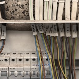 Pogelektro - Otwarta rozdzielnica elektryczna z kolorowymi, opisanymi przewodami (np. 'INDUKCJA', 'Kuchnia', 'Salon') podłączonymi do wyłączników EATON, na tle surowej ściany.