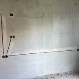 Pogelektro - Szara ściana z betonu z wyciętymi bruzdami pod okablowanie elektryczne, widoczne trzy puszki instalacyjne i odręczne notatki 'Dok. Indukcja 12cm'.
