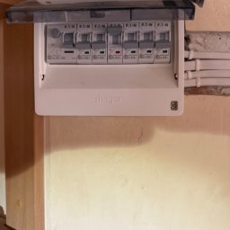 Pogelektro - Biała rozdzielnica elektryczna Shager z sześcioma wyłącznikami nadprądowymi Eaton, zamontowana na beżowej ścianie, z widocznymi przewodami i białymi rurkami instalacyjnymi.