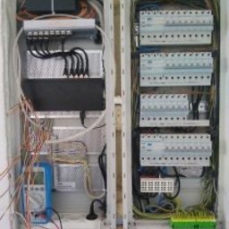 Pogelektro - Otwarta, dwusekcyjna rozdzielnica elektryczna z rzędami wyłączników nadprądowych, uporządkowanym okablowaniem i leżącym miernikiem na dole, w tle nieotynkowana ściana.