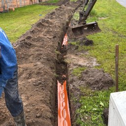 Pogelektro - Wykopany rów z ułożonym pomarańczowym kanałem kablowym, częściowo zasypany ziemią, obok koparka i pracownik w kaloszach, w tle budowany dom z cegły pod pochmurnym niebem.