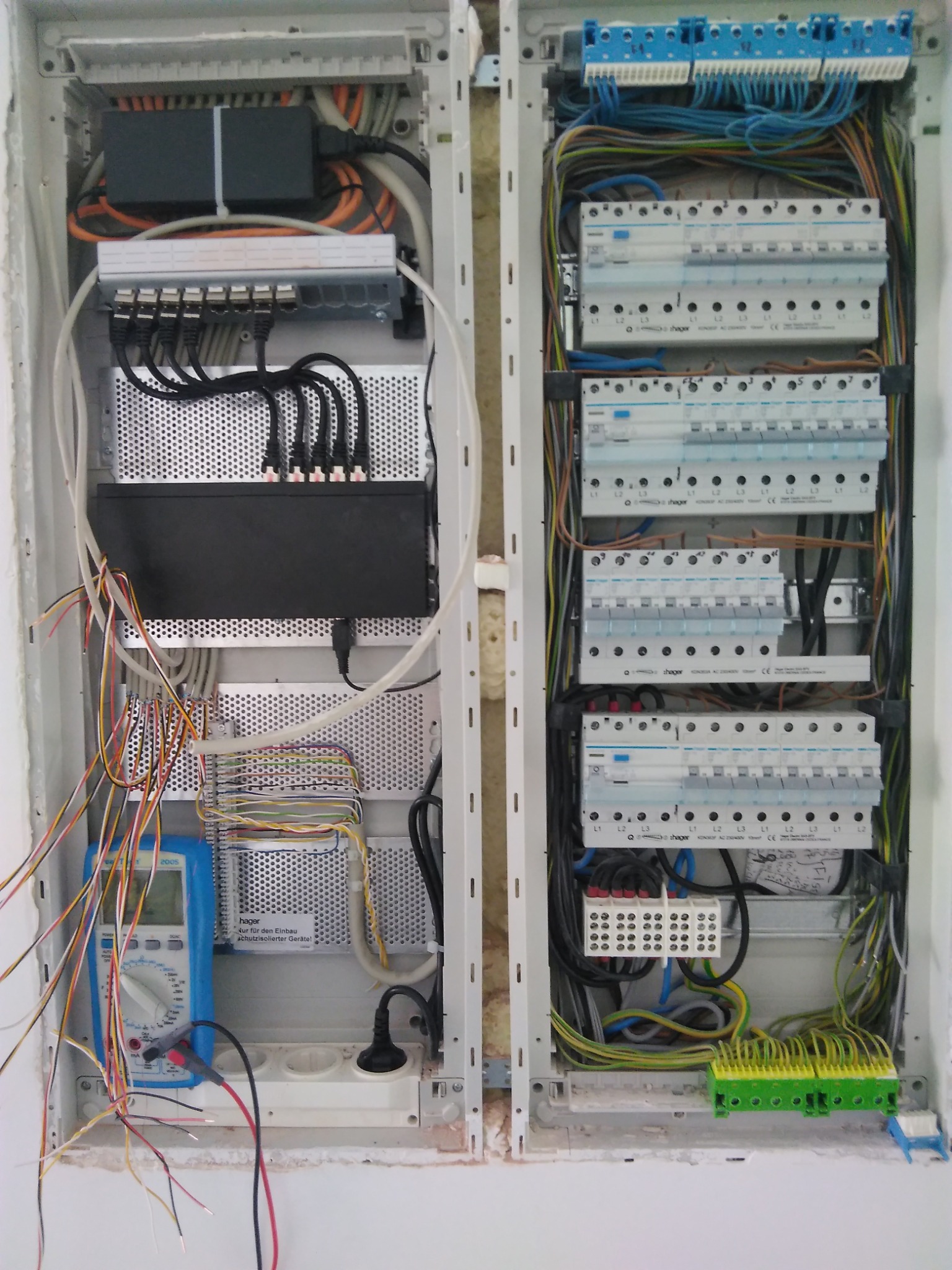 Otwarta szafa rozdzielcza z widocznymi bezpiecznikami, przewodami i urządzeniami elektrycznymi. Po lewej stronie widoczny multimetr z podłączonymi przewodami pomiarowymi. Widoczne także złącza RJ45...