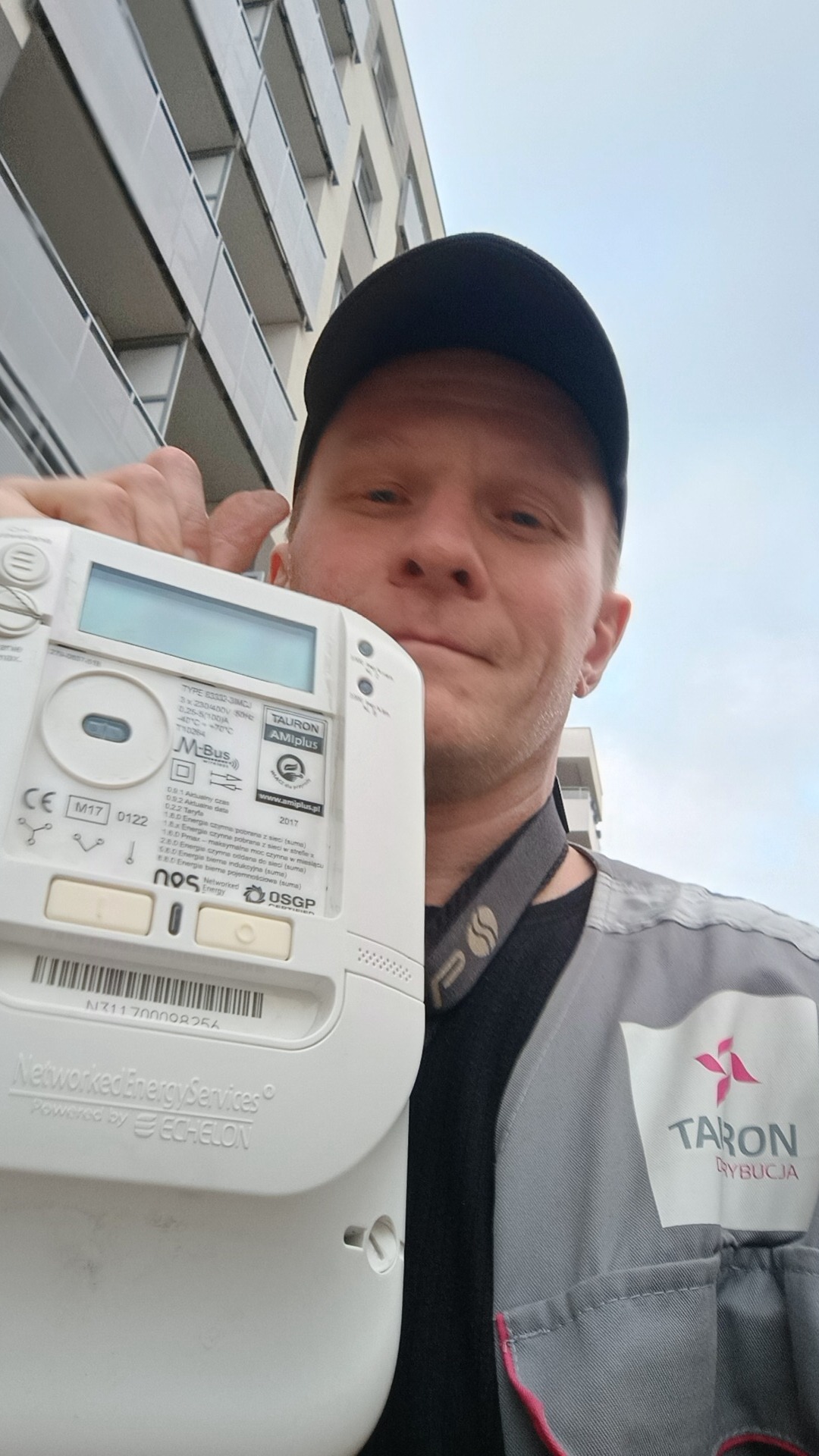 Elektryk w uniformie firmy Taron Rybnicja, trzymający licznik energii Tauron AMIplus na tle budynku mieszkalnego.