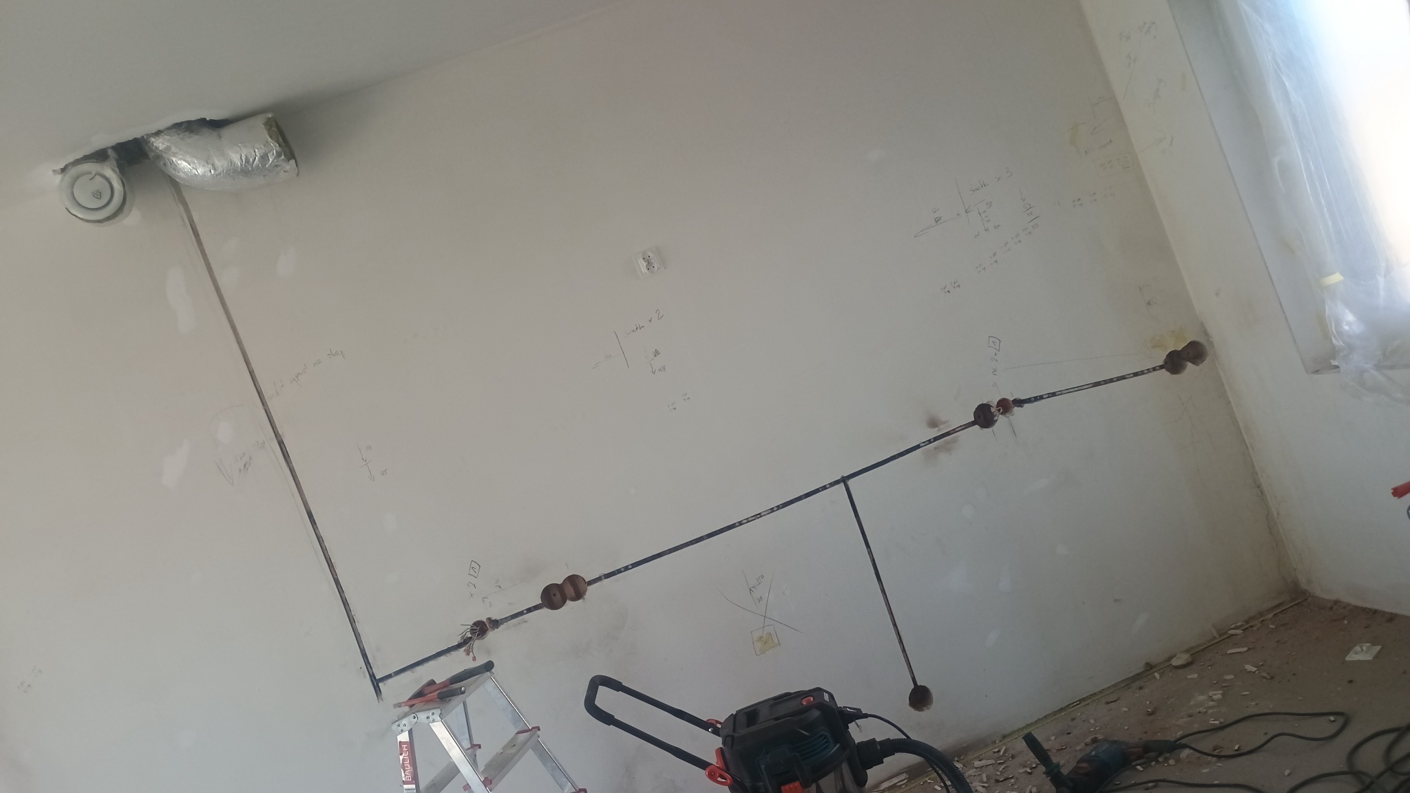 Wnętrze w trakcie instalacji elektrycznej: widoczne przewody poprowadzone po ścianie, oznaczenia na ścianach, drabina i odkurzacz przemysłowy.