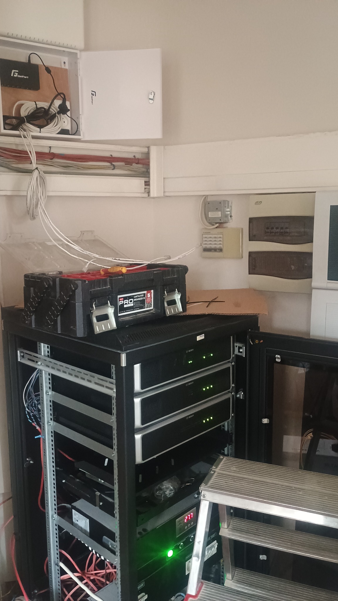 Rozdzielnica elektryczna z otwartą klapą, skrzynka narzędziowa PRO na szafie rack z urządzeniami elektronicznymi i drabina aluminiowa w pomieszczeniu.