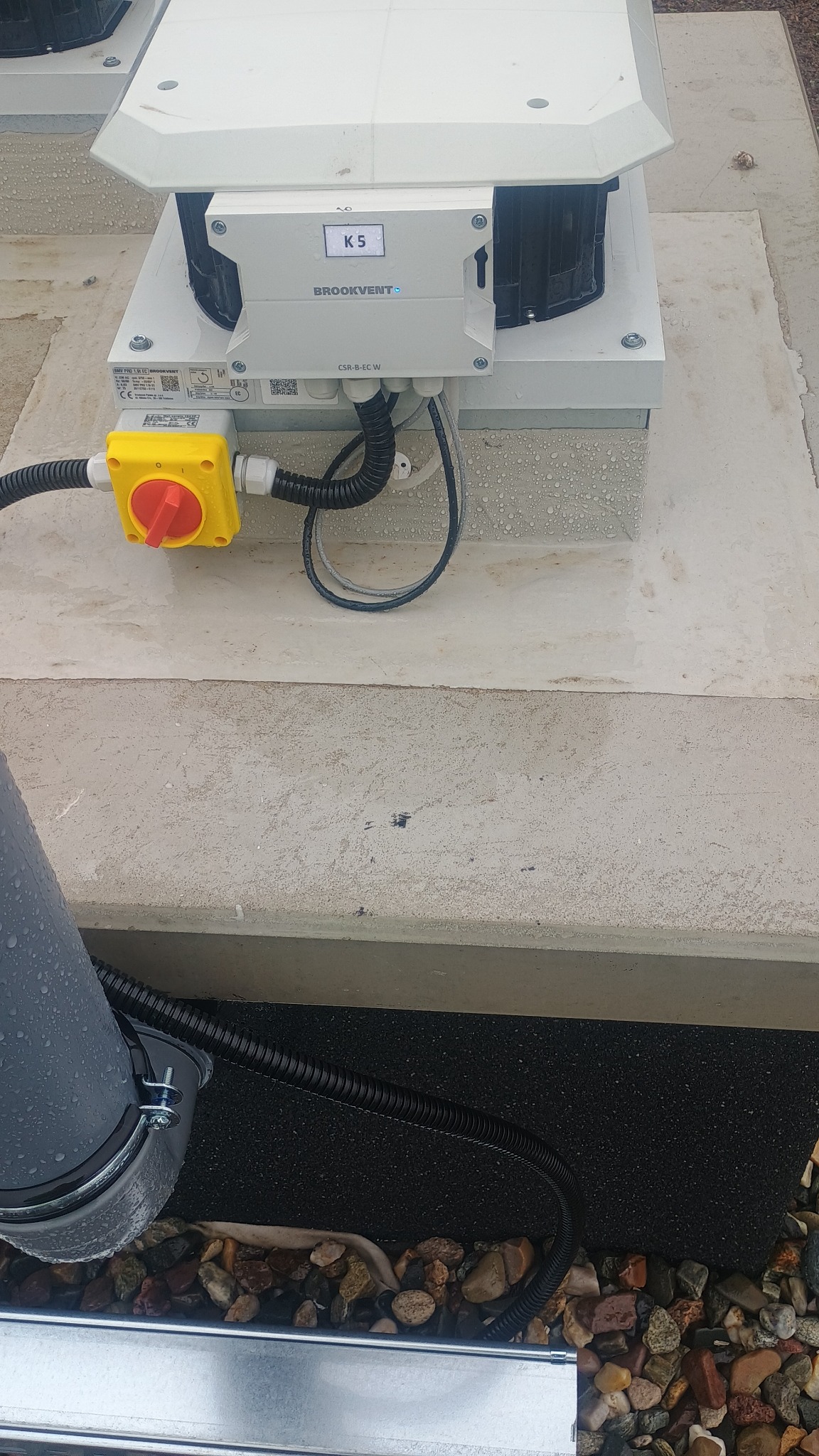 Widok z góry na zainstalowany wentylator dachowy Brookvent CSR-B-EC-W z żółtym wyłącznikiem, połączony przewodami w peszlach, na betonowej podstawie z widocznymi kroplami wody.