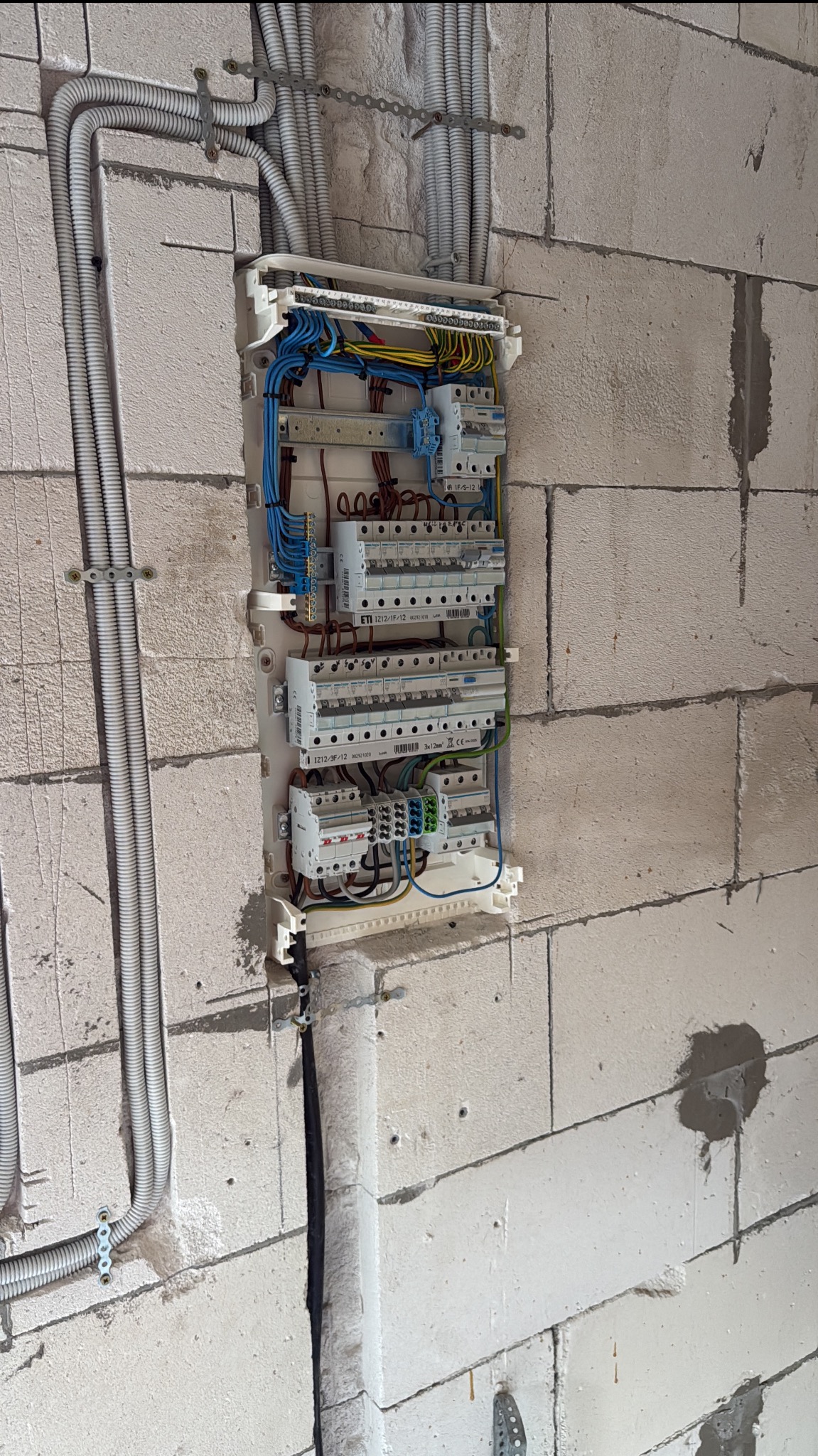 Rozdzielnia elektryczna z przewodami w ścianie z betonu komórkowego. Widoczne zabezpieczenia, kable w peszlach i listwa zaciskowa. Instalacja w trakcie montażu.