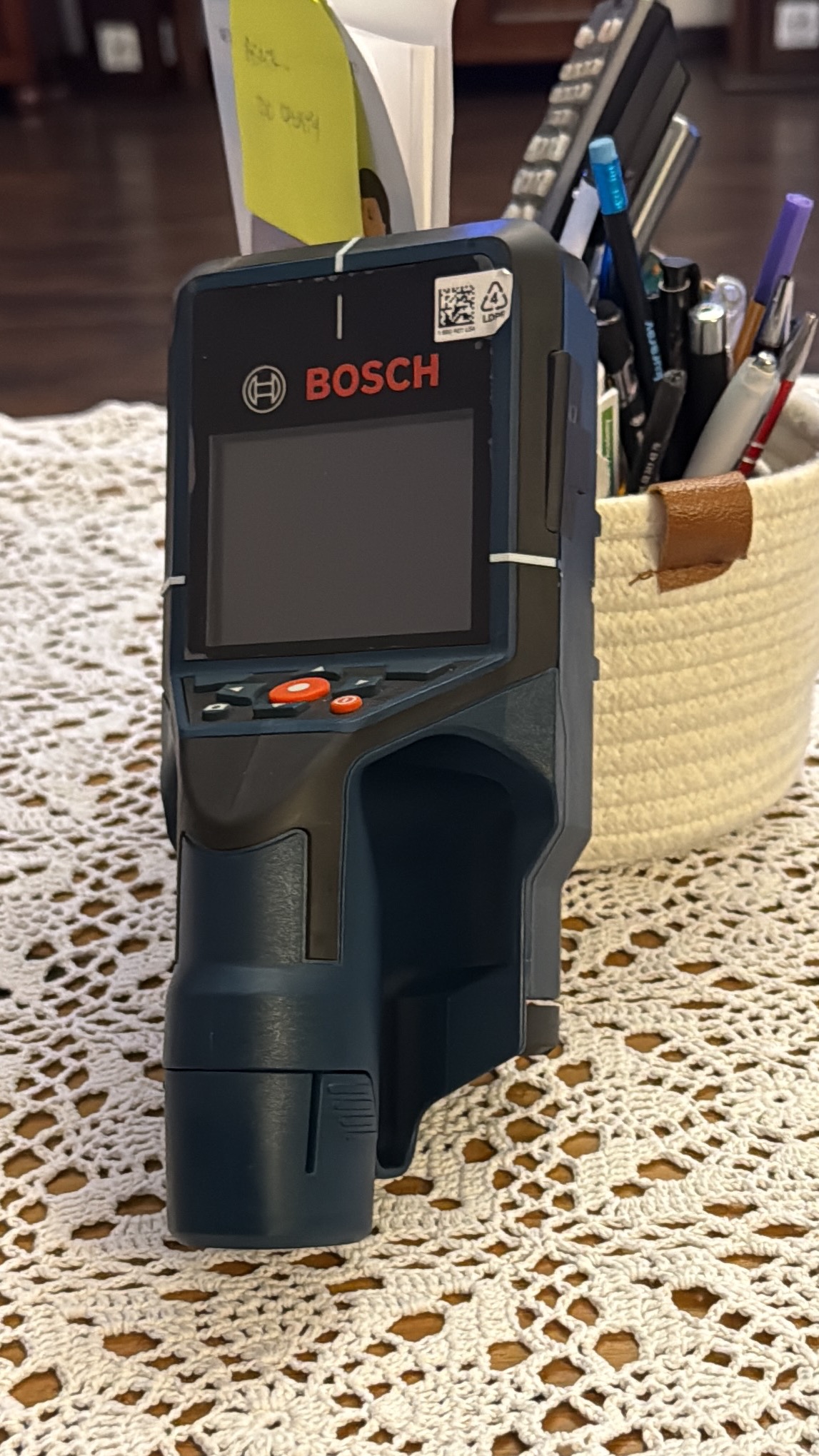 Niwelator laserowy Bosch na stole z haftowanym obrusem, obok koszyka z długopisami i notatkami. Sprzęt pomiarowy dla elektryka, używany do precyzyjnych pomiarów.