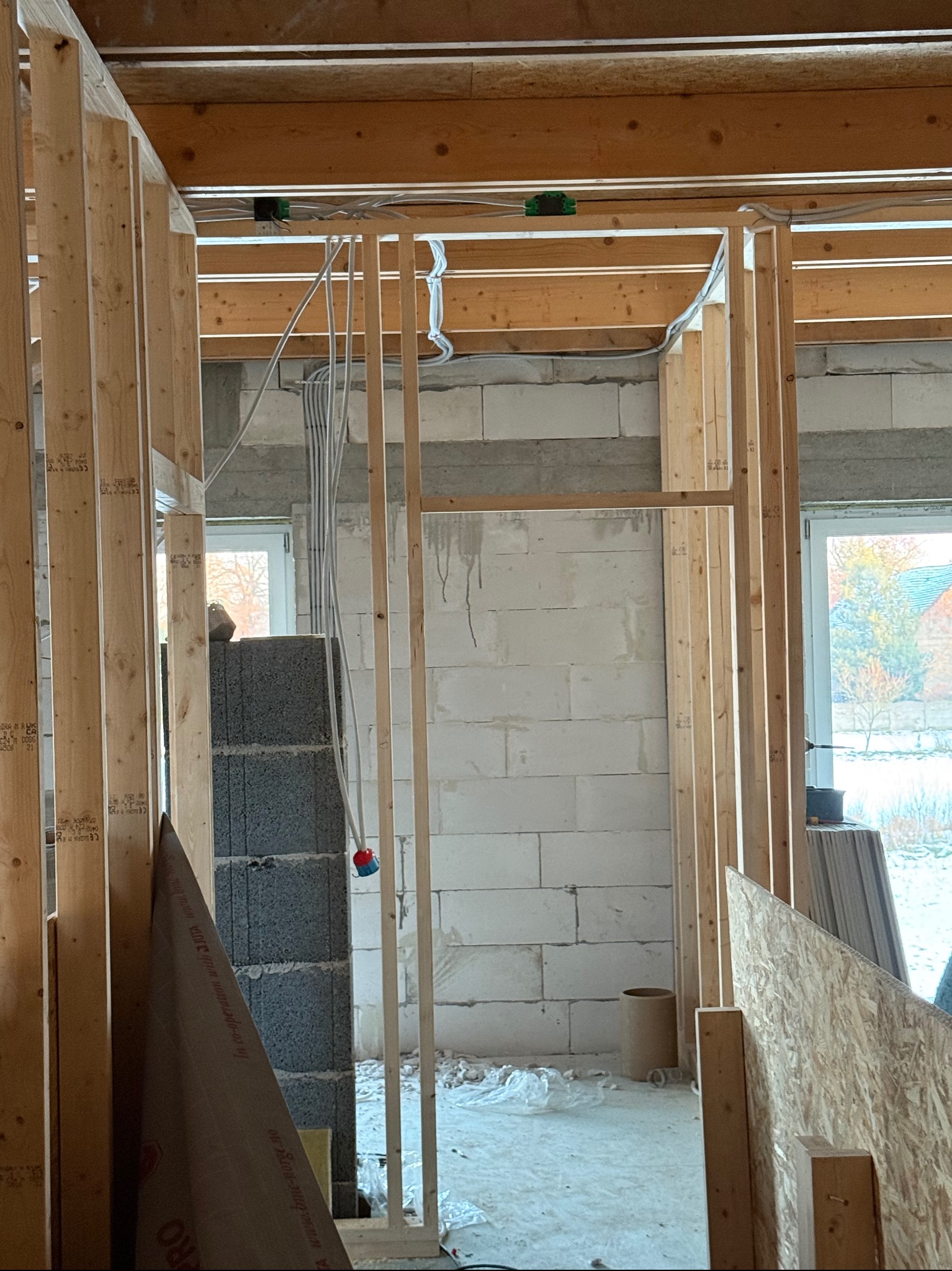 Wnętrze w trakcie budowy z widoczną drewnianą konstrukcją szkieletową i przewodami elektrycznymi poprowadzonymi pod sufitem. Widoczne okna i ściany z bloczków.