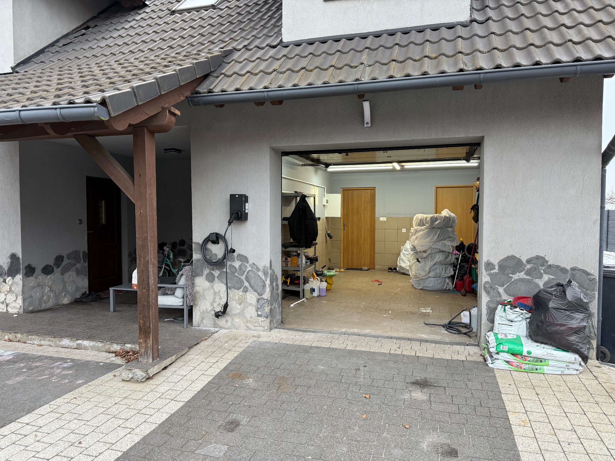 Zewnętrzna ładowarka do samochodu elektrycznego zamontowana na ścianie garażu z dekoracyjnym wykończeniem imitującym kamień. Widoczny kabel i wtyczka.