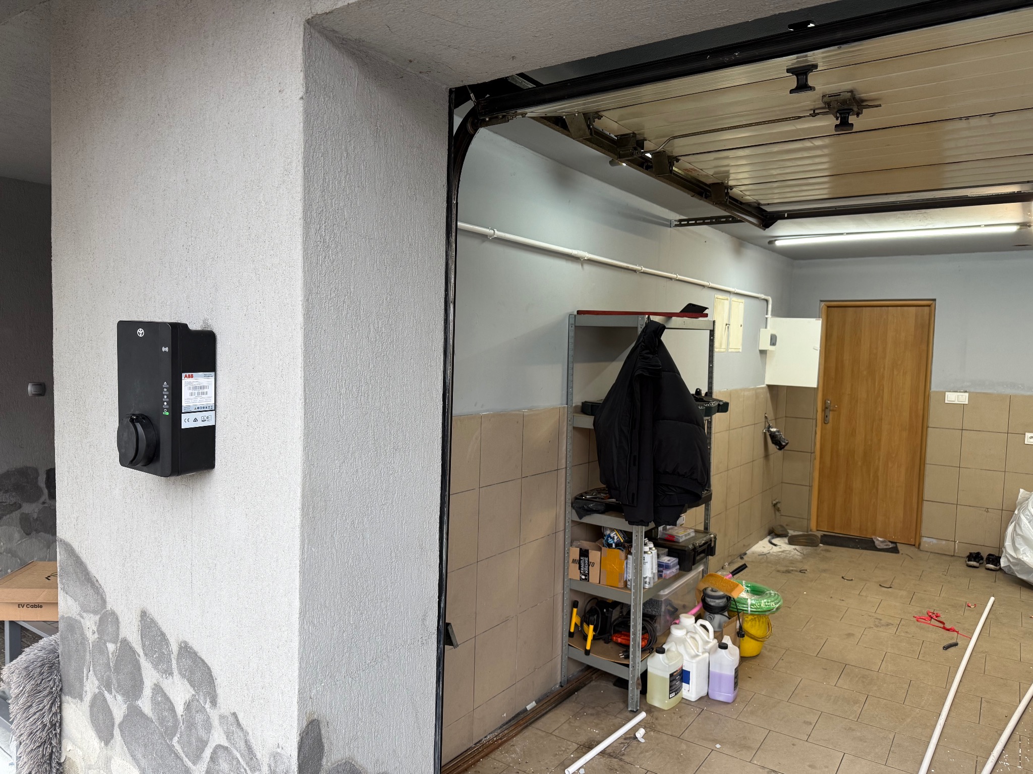 Czarna stacja ładowania EV marki ABB zamontowana na szarej, teksturowanej ścianie garażu z otwartą bramą segmentową i wyposażeniem warsztatowym w tle.