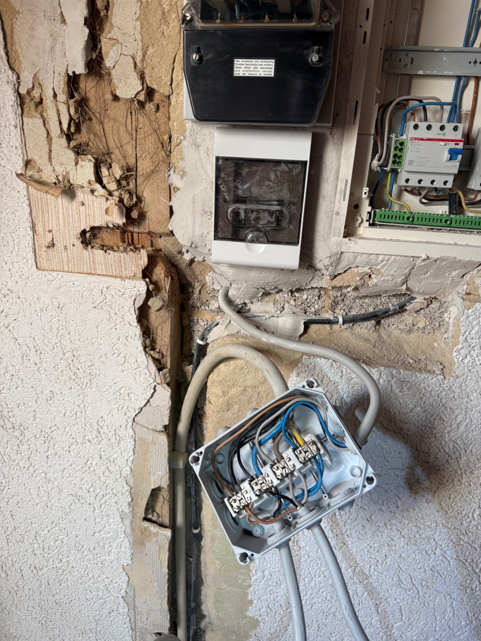 Otwarta puszka elektryczna z przewodami na tle uszkodzonej ściany, widoczny licznik i skrzynka z bezpiecznikami. Prace instalacyjne w trakcie remontu.