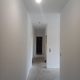 Budimar Bartosz Lewandowski - Korytarz w trakcie malowania, ściany świeżo pomalowane na biało, surowa podłoga, widoczne gniazdka elektryczne i włączniki, goła żarówka.