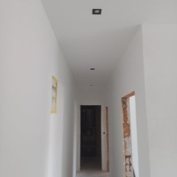 Budimar Bartosz Lewandowski - Korytarz po malowaniu na biało, widoczne czarne, kwadratowe lampy sufitowe, fragment zabezpieczonej taśmą malarską ściany i surowe wykończenie ościeżnicy.