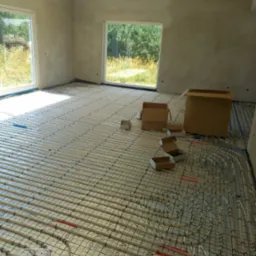 Rozłożona instalacja ogrzewania podłogowego w surowym wnętrzu, widoczne kartony i okna z widokiem na zieleń.