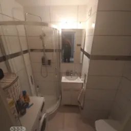 Ukończona łazienka z kabiną prysznicową, umywalką z szafką, toaletą i pralką, wykończona białymi płytkami z ciemnym akcentem. Widoczne odbicie osoby robiącej zdjęcie w lustrze.