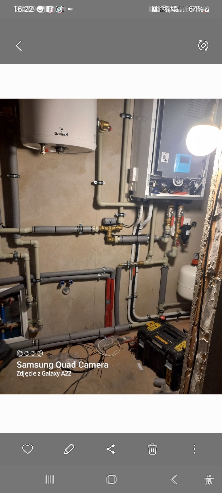 Złożona instalacja grzewcza z kotłem i zasobnikiem Galmet, rury z izolacją, narzędzia DeWalt na podłodze, widok na surową ścianę w trakcie remontu.