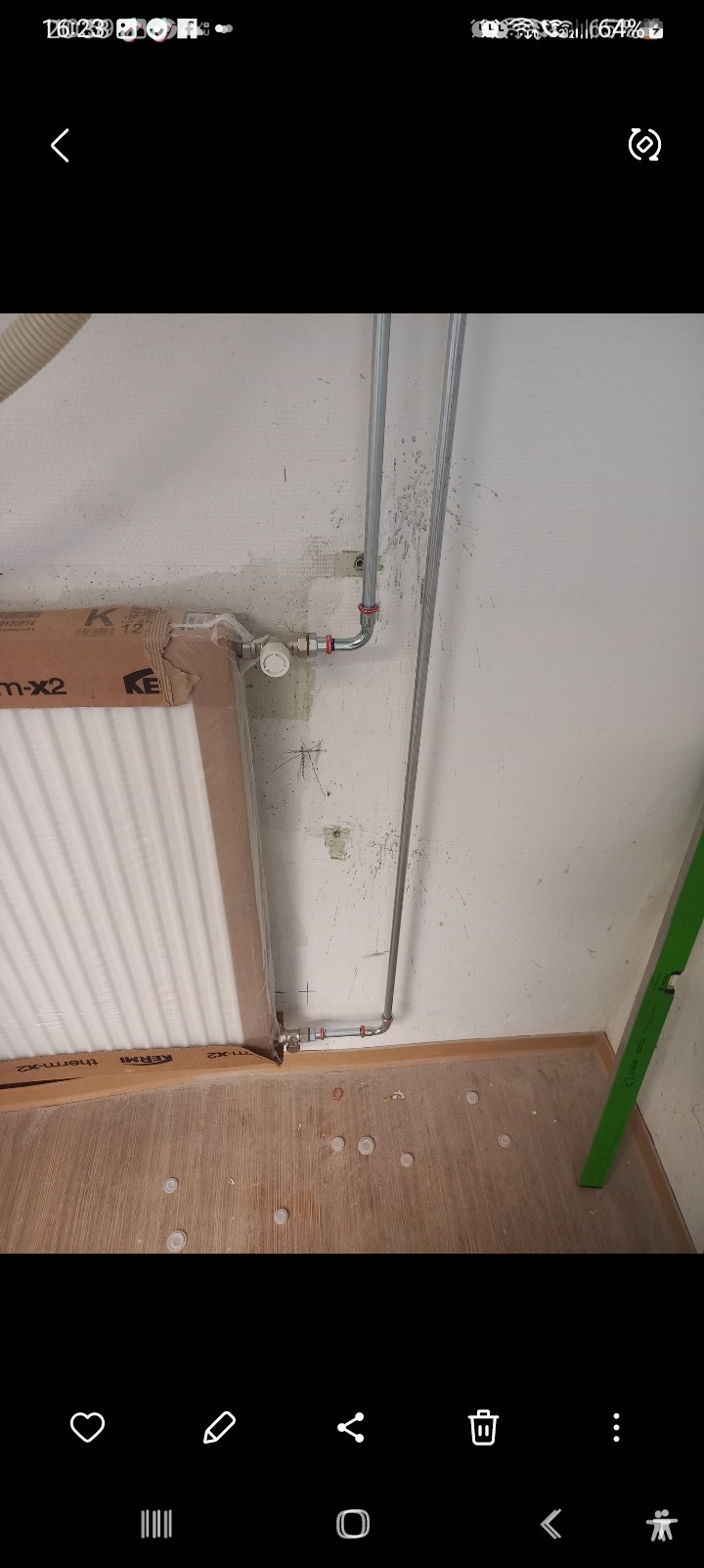 Niezamontowany grzejnik marki KERMI w opakowaniu, rury instalacji centralnego ogrzewania z zaworem termostatycznym przy ścianie w trakcie montażu, fragment poziomicy na podłodze z nakrętkami...