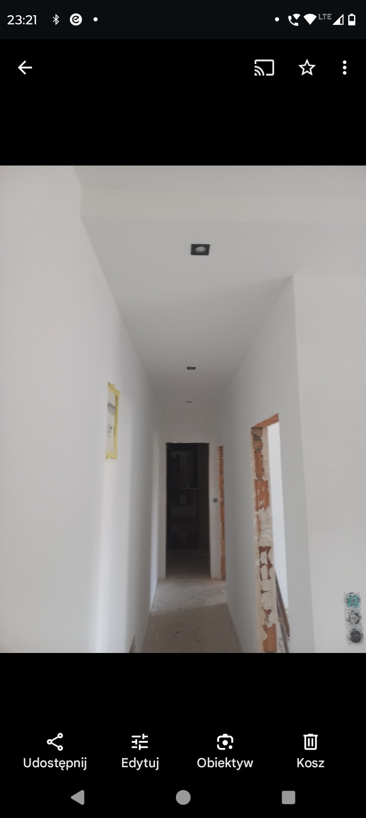 Korytarz po malowaniu na biało, widoczne czarne, kwadratowe lampy sufitowe, fragment zabezpieczonej taśmą malarską ściany i surowe wykończenie ościeżnicy.