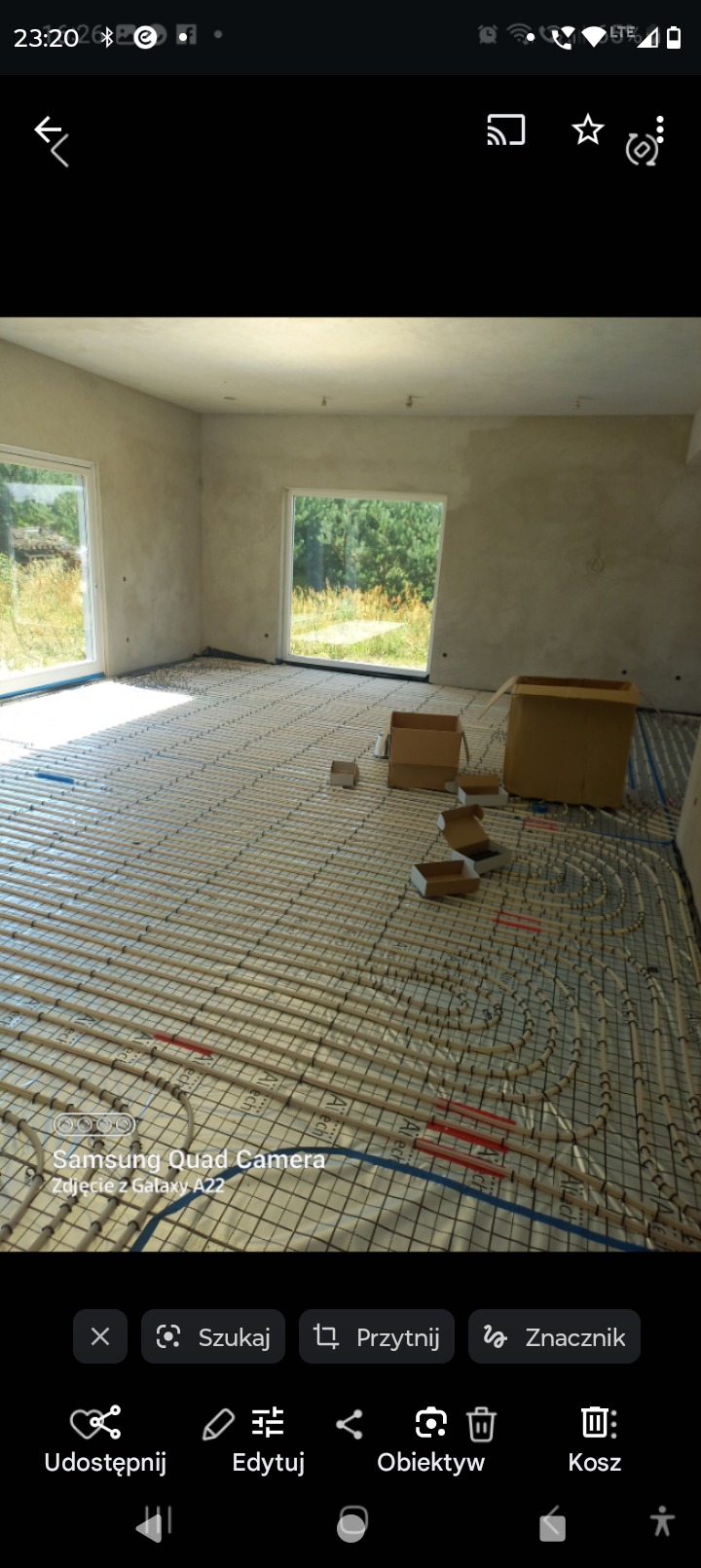 Rozłożona instalacja ogrzewania podłogowego w surowym wnętrzu, widoczne kartony i okna z widokiem na zieleń.