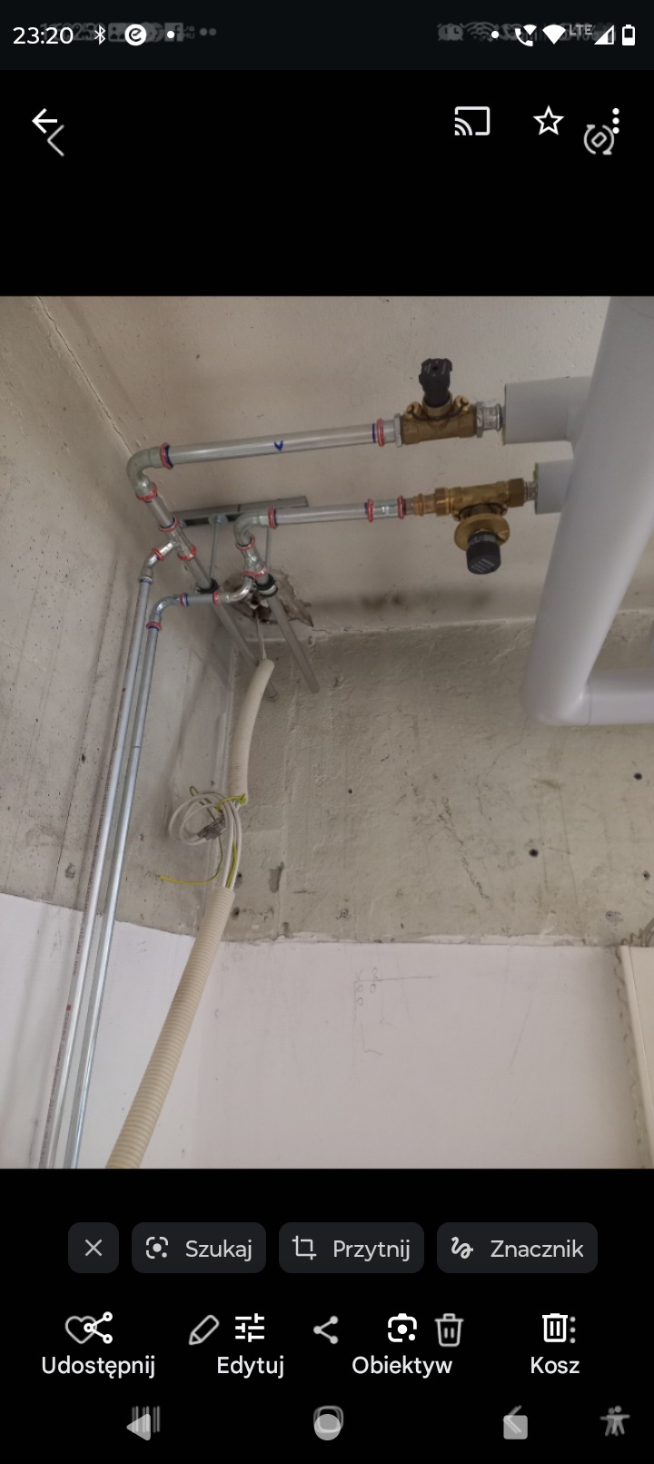 Szare rury instalacji wodnej z zaworami i rurą osłonową kabli elektrycznych na tle surowego, betonowego sufitu i ściany.