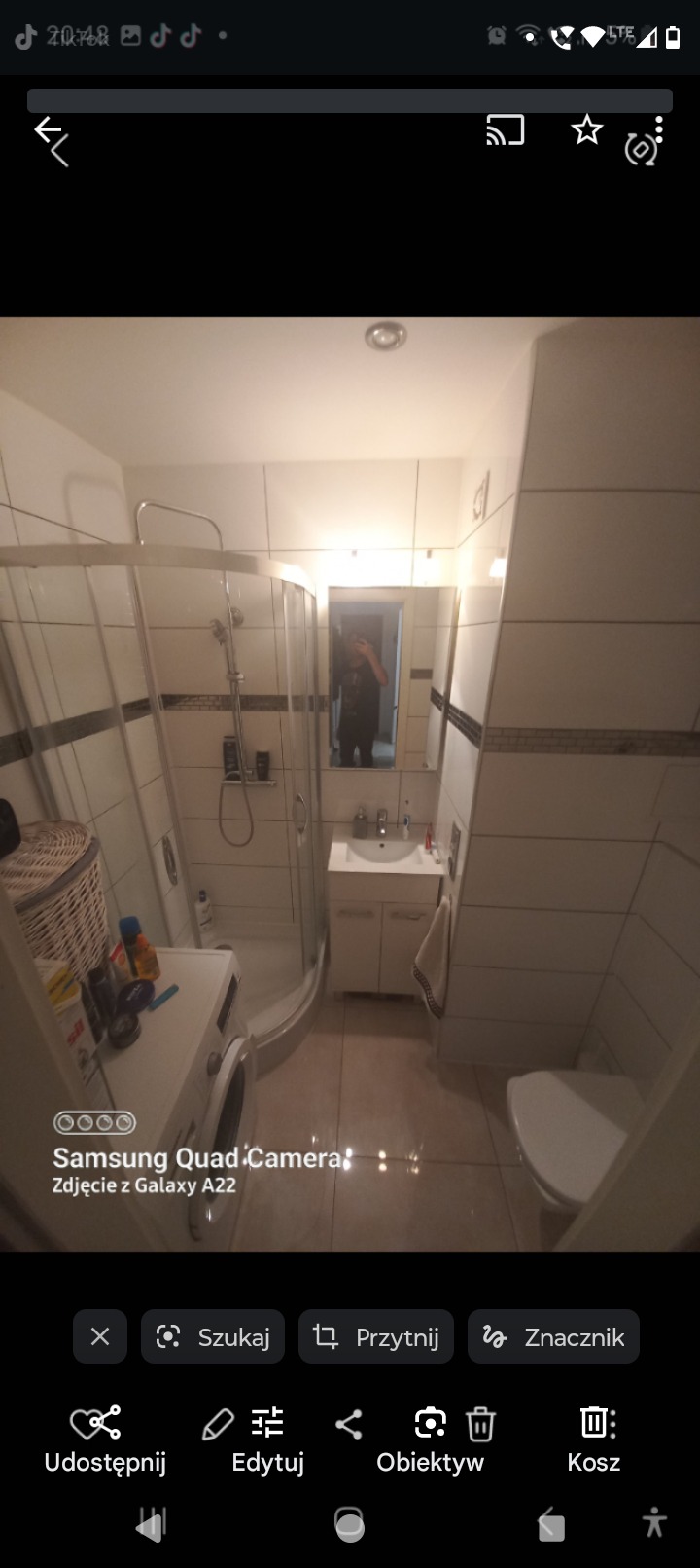 Ukończona łazienka z kabiną prysznicową, umywalką z szafką, toaletą i pralką, wykończona białymi płytkami z ciemnym akcentem. Widoczne odbicie osoby robiącej zdjęcie w lustrze.