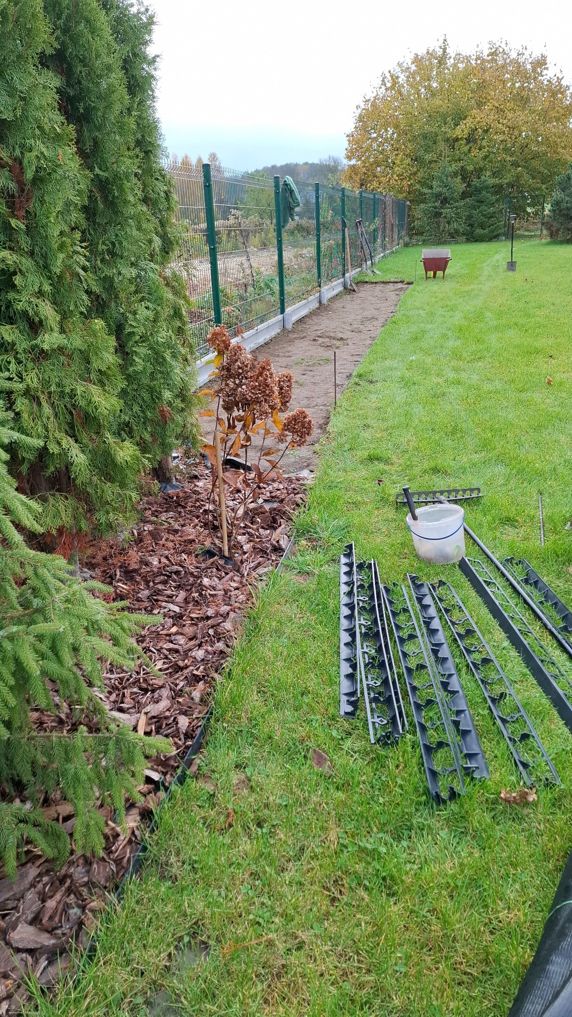 Przygotowania do założenia rabaty w ogrodzie: widoczne obrzeża trawnikowe, kora, narzędzia ogrodnicze i wytyczona ścieżka z ziemi wzdłuż ogrodzenia.