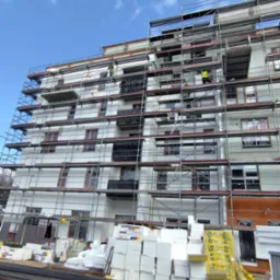 Wielopiętrowy budynek w trakcie ocieplania, otoczony rusztowaniami. Widoczne materiały izolacyjne i pracownicy na rusztowaniu. Błękitne niebo w tle.