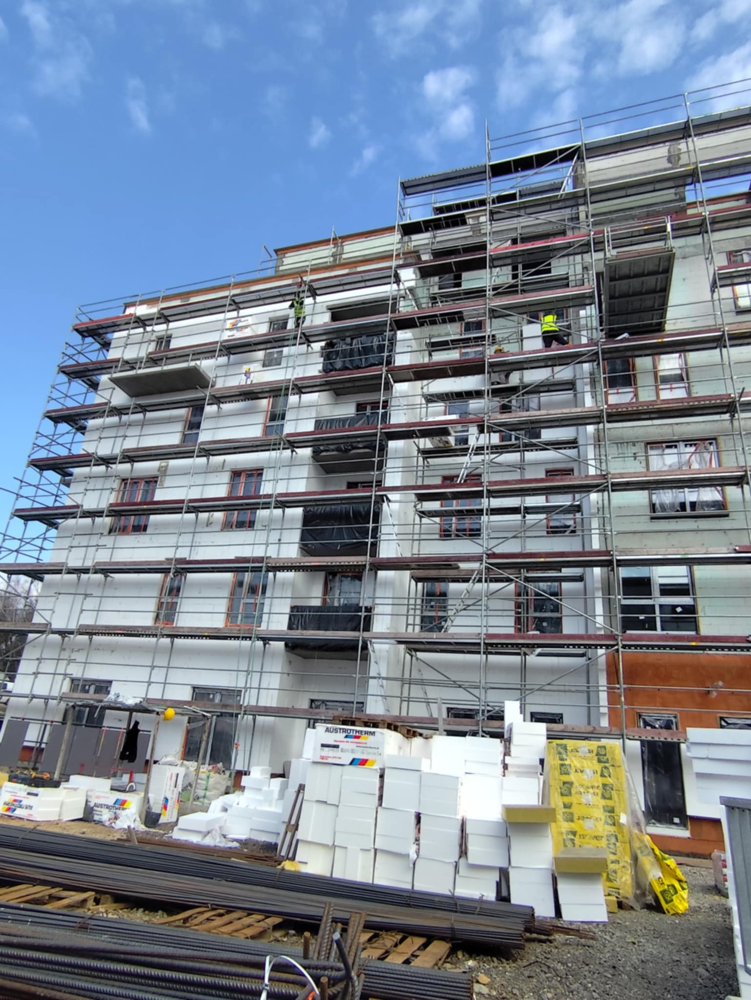 Wielopiętrowy budynek w trakcie ocieplania, otoczony rusztowaniami. Widoczne materiały izolacyjne i pracownicy na rusztowaniu. Błękitne niebo w tle.