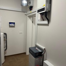 SOLAR ENERGY - Zainstalowany magazyn energii Pylontech obok czerwonej gaśnicy, z falownikiem na ścianie i otwartą skrzynką elektryczną powyżej drzwi w pomieszczeniu technicznym.