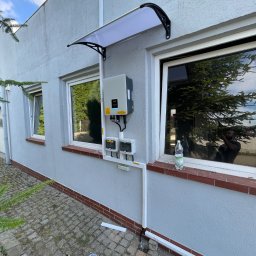 SOLAR ENERGY - Montaż instalacji fotowoltaicznej na szarej ścianie budynku, widoczny inwerter pod daszkiem nad oknem, zabezpieczenia elektryczne, szlifierka kątowa na kostce brukowej, instalacja w trakcie...