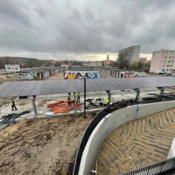 SOLAR ENERGY - Montaż zadaszenia z paneli słonecznych nad parkingiem, widoczne elementy konstrukcyjne, pracownicy w kamizelkach odblaskowych, w tle zabudowa miejska i pochmurne niebo.