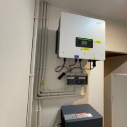 Zamontowany biały falownik Sofar z podłączonymi przewodami, umieszczony na ścianie obok białych rur instalacyjnych i szarego magazynu energii, widoczne zabezpieczenia elektryczne.