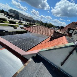 SOLAR ENERGY - Panele słoneczne zamontowane na czerwonym dachu domu jednorodzinnego, widok z góry, w tle błękitne niebo z chmurami i budynek hali sportowej.