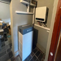 SOLAR ENERGY - Biały magazyn energii Pylontech z szarym blatem, zamontowany w rogu pomieszczenia z szarą ścianą i brązowymi drzwiami, obok widać biały inwerter z podłączonymi kablami.