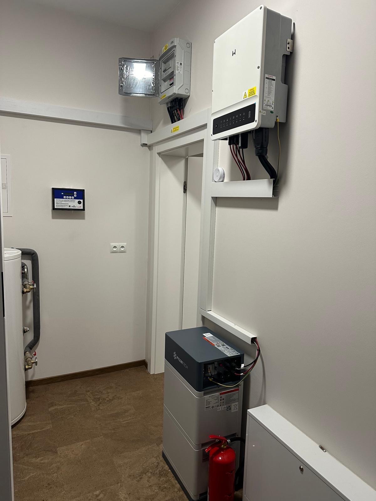 Zainstalowany magazyn energii Pylontech obok czerwonej gaśnicy, z falownikiem na ścianie i otwartą skrzynką elektryczną powyżej drzwi w pomieszczeniu technicznym.