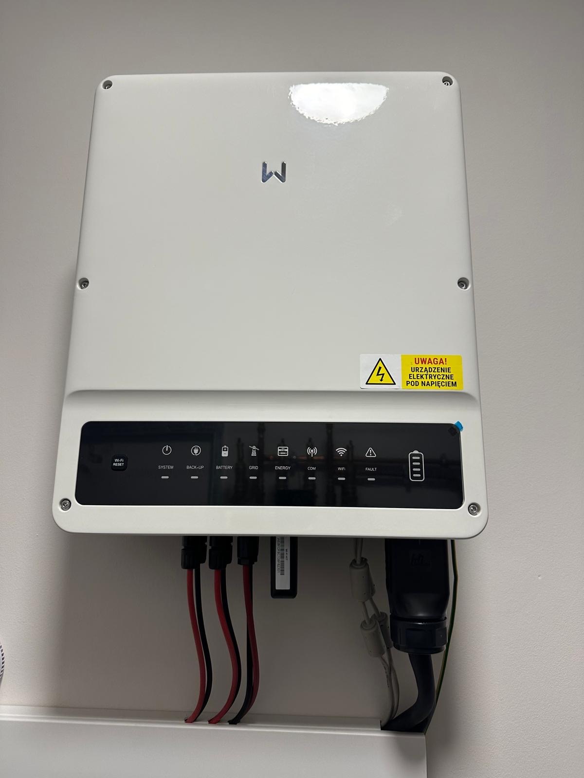 Biała obudowa inwertera z widocznymi złączami kablowymi, diodami sygnalizacyjnymi i naklejką ostrzegawczą o napięciu elektrycznym.