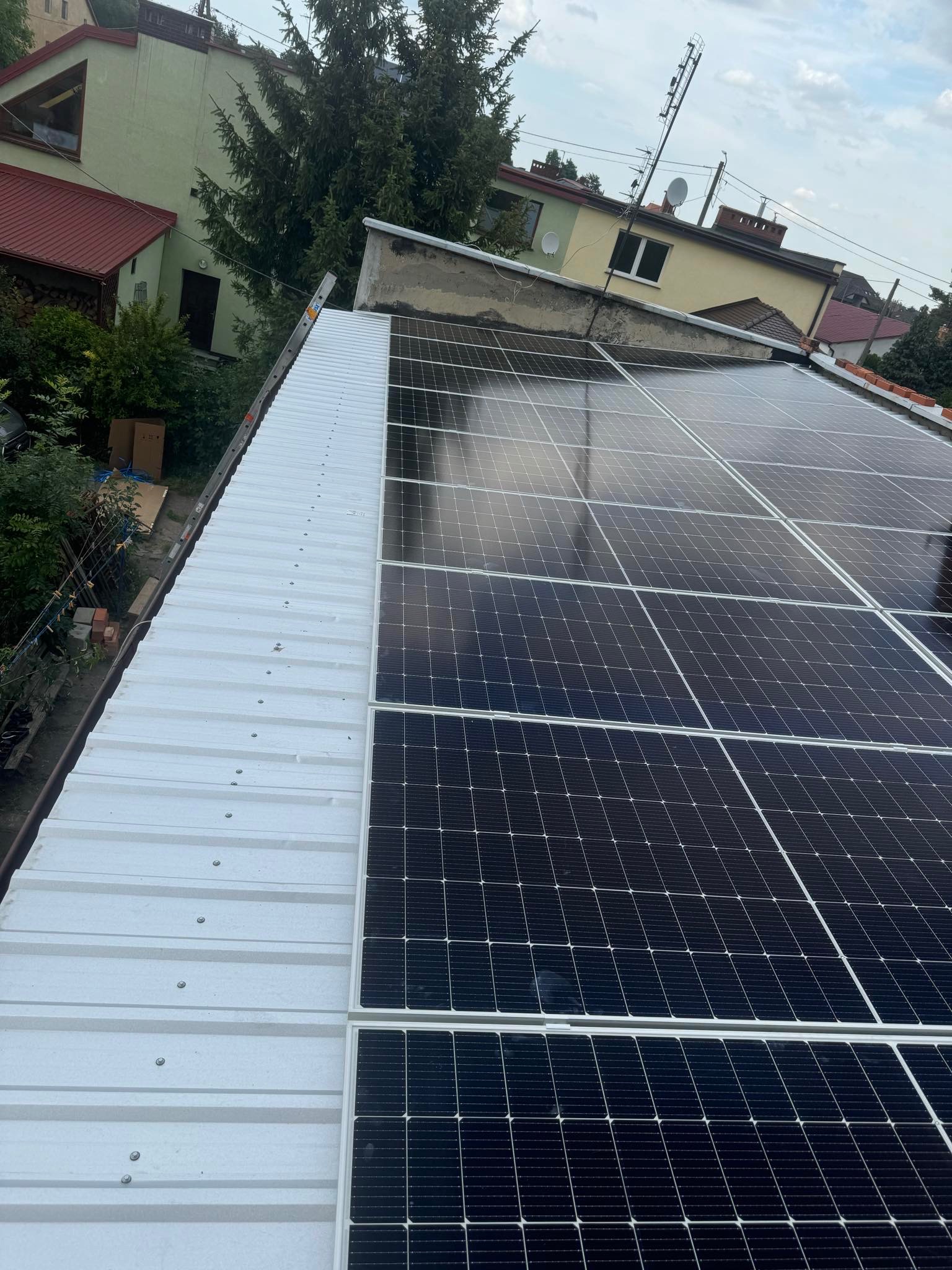 Panel fotowoltaiczny zamontowany na białym, metalowym dachu, widok z góry, w tle budynki mieszkalne i drzewa.