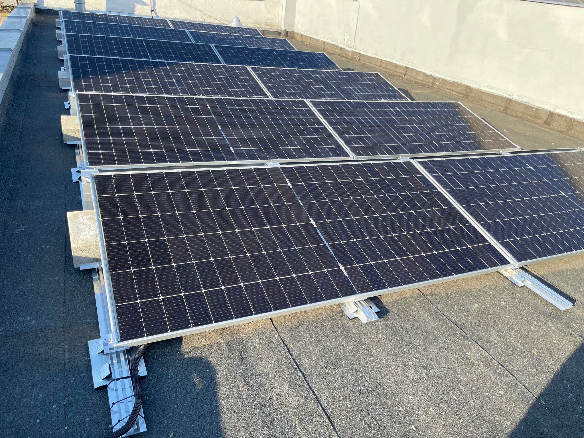 Rząd paneli fotowoltaicznych na płaskim dachu pokrytym papą, widoczne aluminiowe mocowania i peszel z kablami.