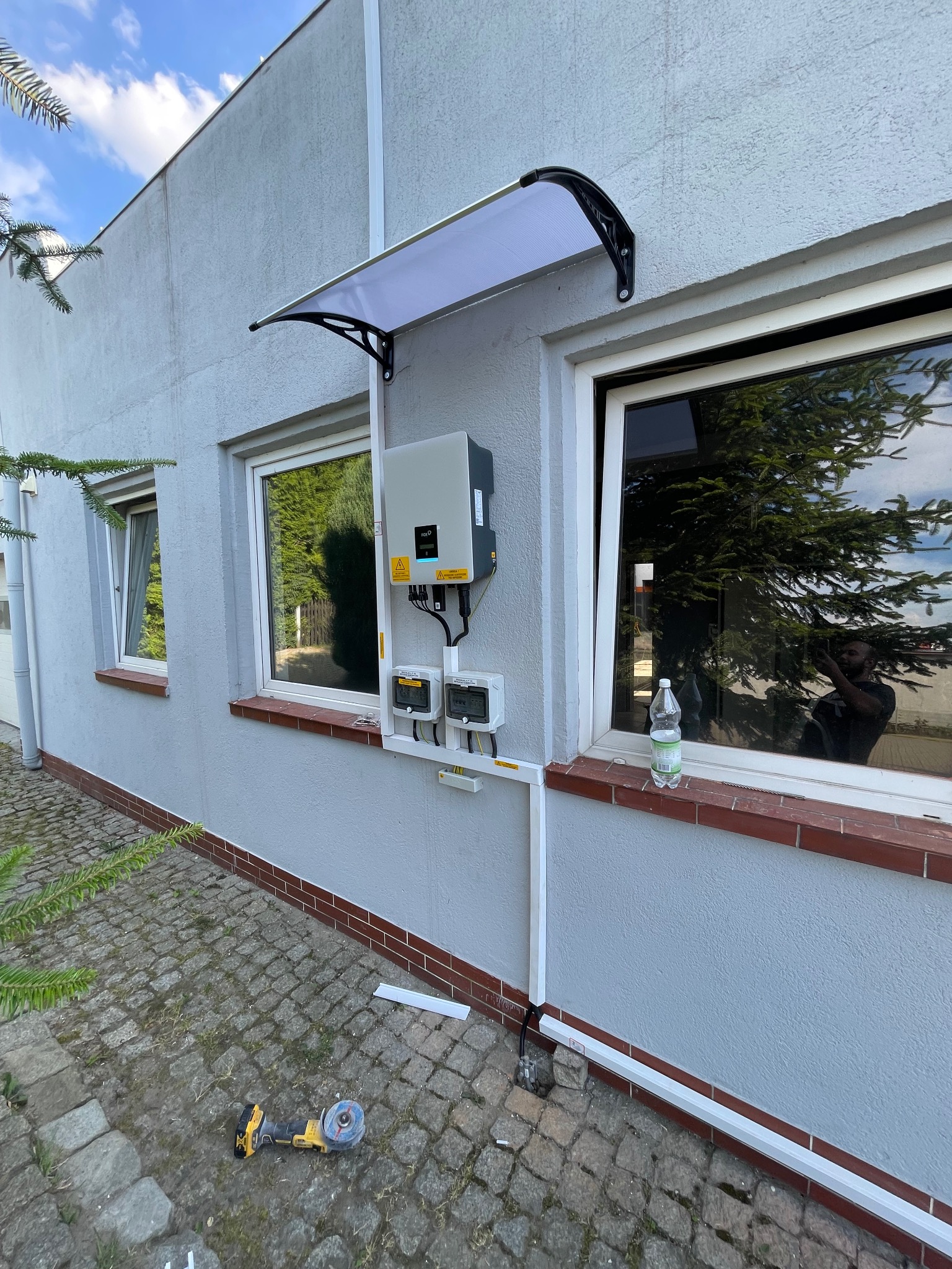 Montaż instalacji fotowoltaicznej na szarej ścianie budynku, widoczny inwerter pod daszkiem nad oknem, zabezpieczenia elektryczne, szlifierka kątowa na kostce brukowej, instalacja w trakcie...
