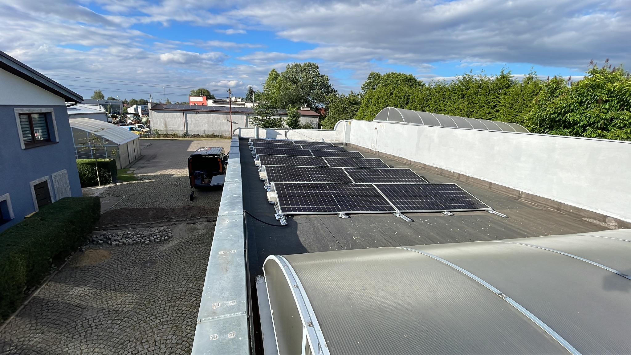 Rząd paneli słonecznych na płaskim dachu budynku przemysłowego, z widocznym samochodem dostawczym i fragmentem brukowanego placu w tle, przy słonecznej pogodzie.