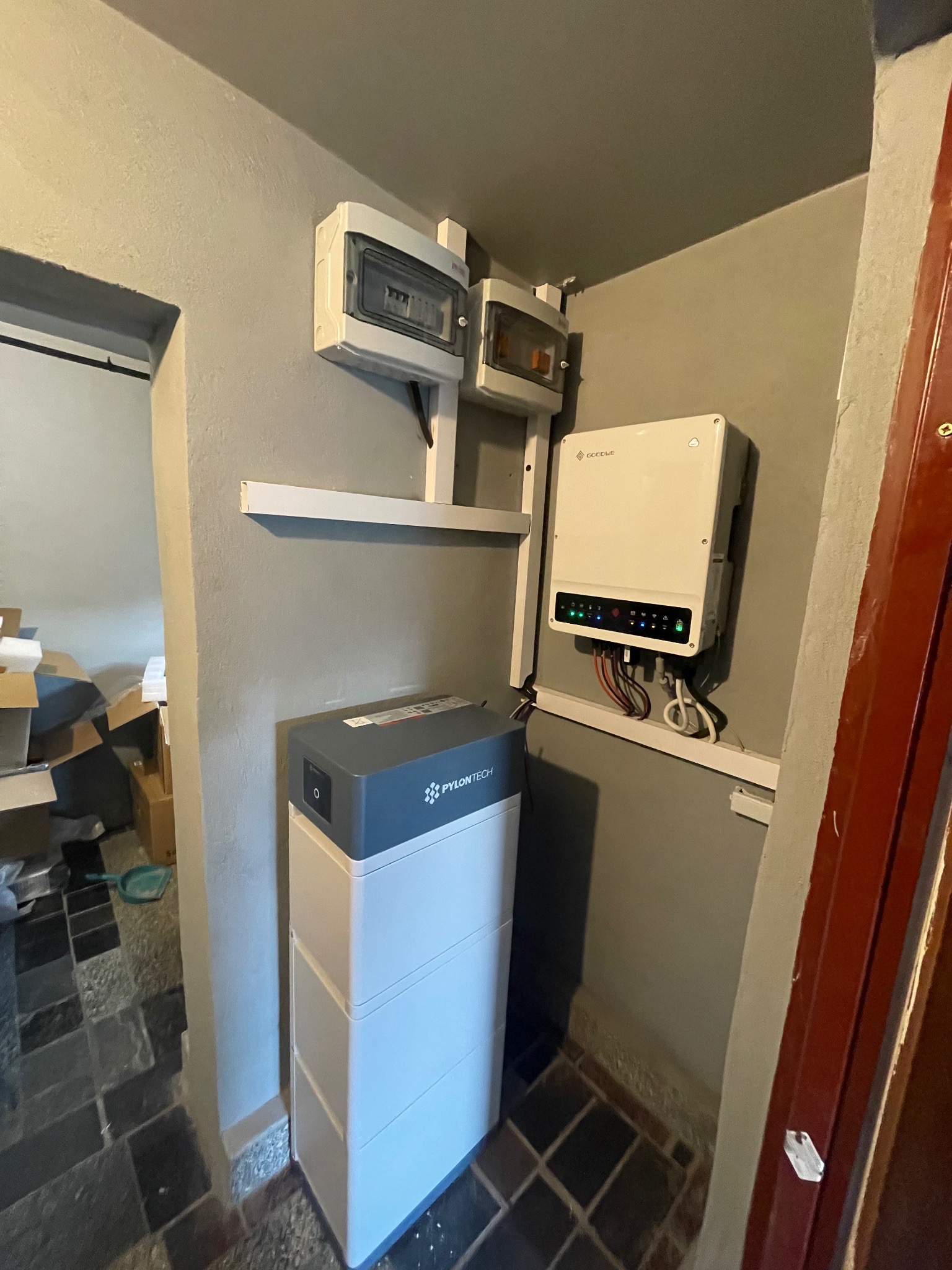 Wnętrze pomieszczenia z zamontowanym magazynem energii Pylontech, inwerterem Goodwe z diodami LED oraz dwoma skrzynkami elektrycznymi.
