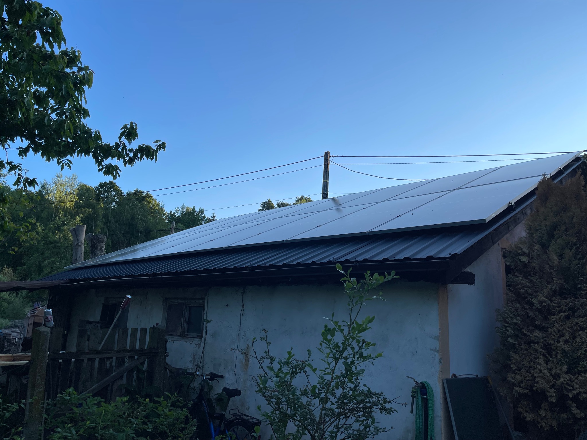 Panele słoneczne na dachu starszego, otynkowanego budynku gospodarczego z widocznym ogrodem i niebieskim niebem w tle.