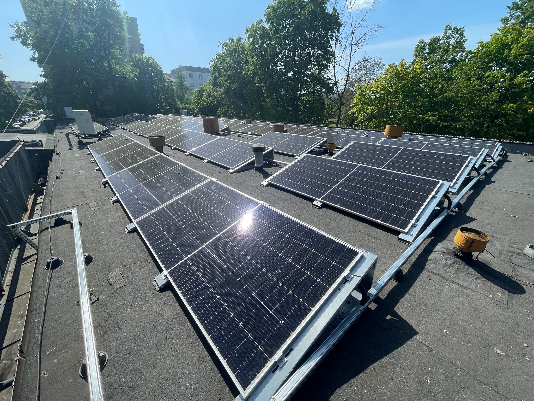 Rząd paneli słonecznych z odbiciami słońca, zamontowanych na płaskim dachu budynku z widocznymi kominami wentylacyjnymi i zielonymi drzewami w tle.