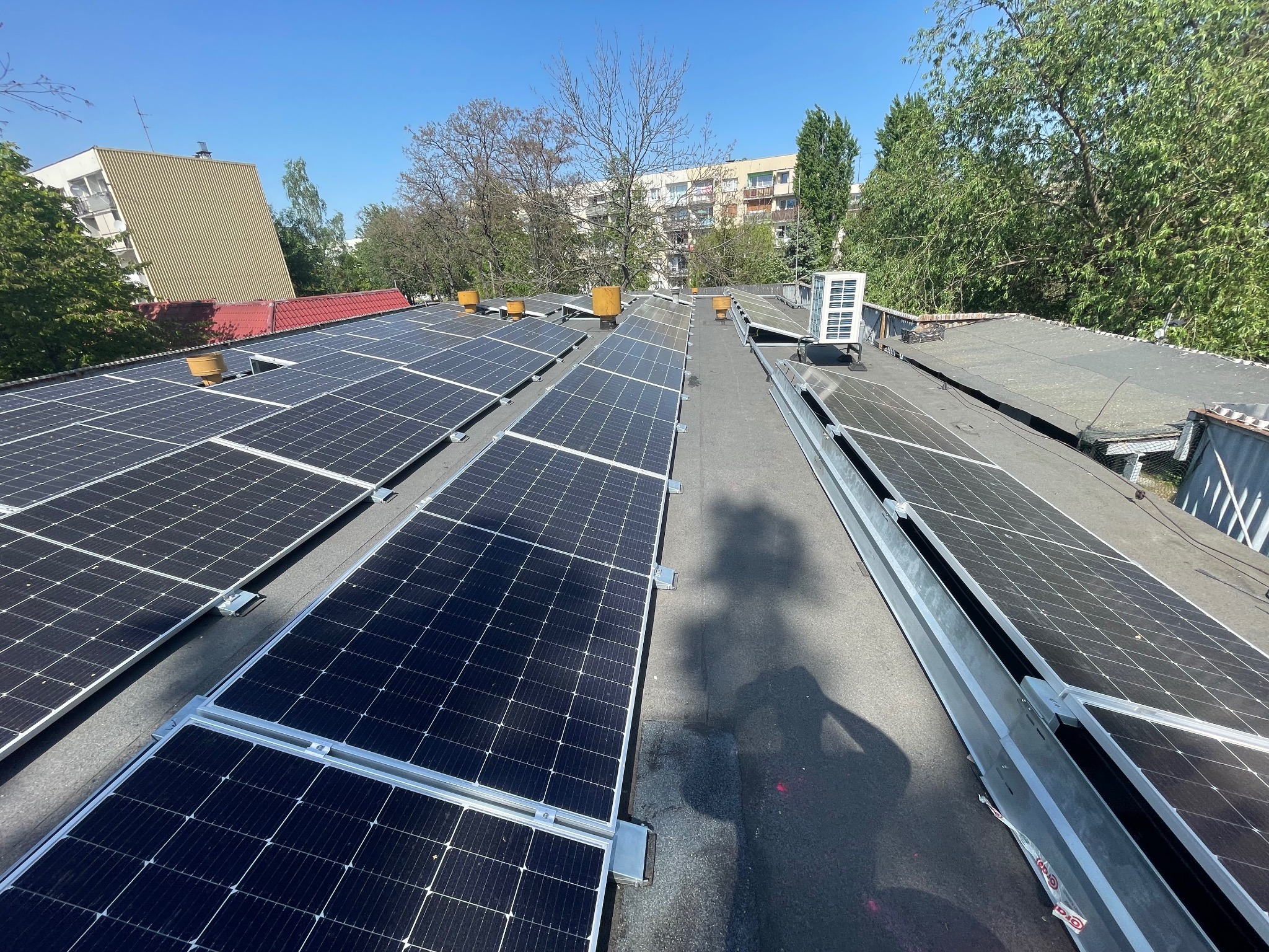 Widok z góry na instalację paneli słonecznych na płaskim dachu budynku z widocznymi elementami wentylacyjnymi i klimatyzatorem, na tle drzew i budynków mieszkalnych.