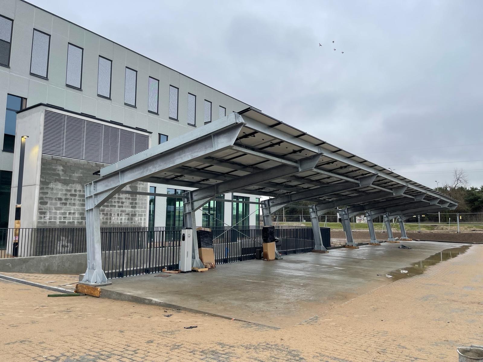 Zadaszenie parkingu z panelami fotowoltaicznymi na metalowej konstrukcji, w tle nowoczesny budynek z rzędem okien, teren budowy z kałużami i piaskiem, pochmurne niebo z przelatującymi ptakami.